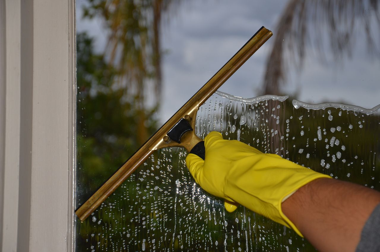 services-04 A close-up view of a hand cleaning an outdoor window with a squeegee while wearing yellow gloves.
